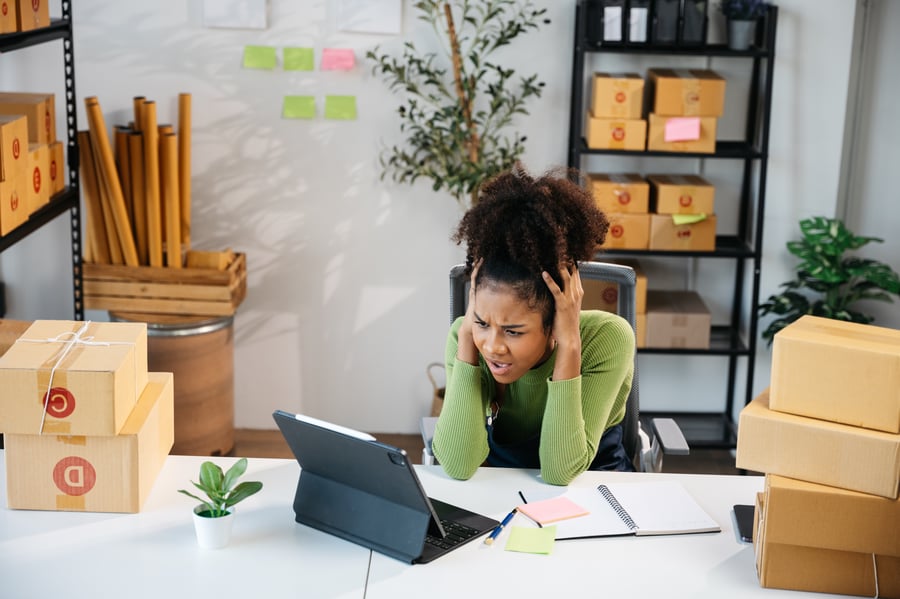 A woman surrounded by boxes looks upset while looking at a tablet at her desk