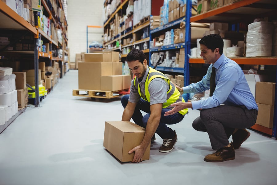 A man teaching another man how to lift a heavy box in a warehouse