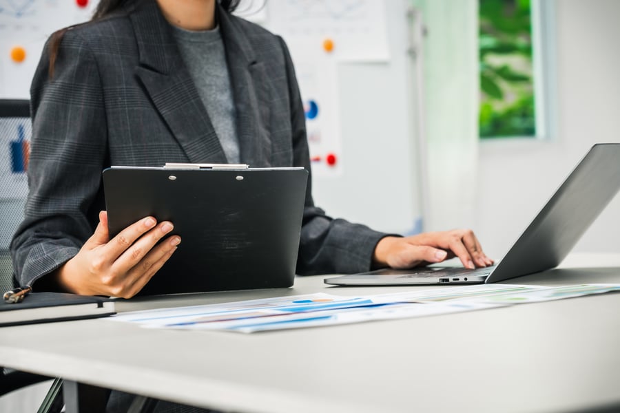 A woman looks at a clipboard and a laptop in an office